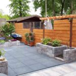 Elegante tuin met drie comfortabele tuinstoelen rond een stijlvolle tafel op een fraai bestraat oppervlak met mooie houten hekken en stenen baken voor de planten